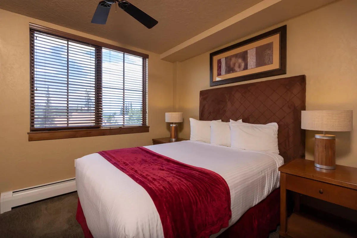 A comfortable bedroom with a bed dressed in a red throw blanket, window with blinds, and decorative artwork on the walls. The simple, soothing color palette and soft lighting evoke a sense of calm.