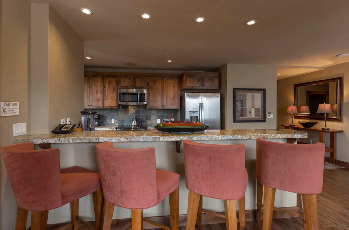 A bright, inviting kitchen and breakfast bar setup, accented by granite countertops, bar stools for casual dining, and visible modern appliances. The warm lighting and functional design encourage social gatherings and shared meals.