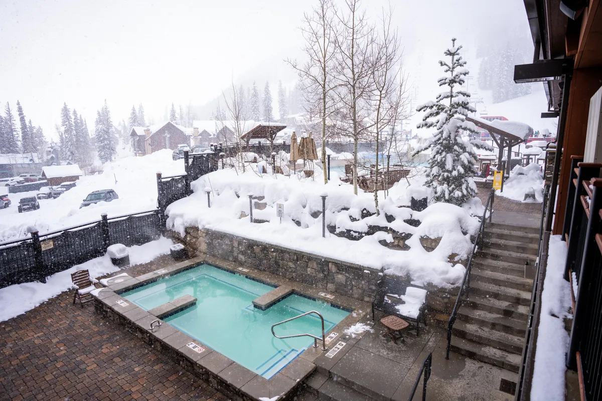 An outdoor hot tub surrounded by snowy landscape and trees, with inviting steam rising from the water. Stone steps lead to the hot tub, creating a welcoming scene for après-ski relaxation amidst winter beauty.