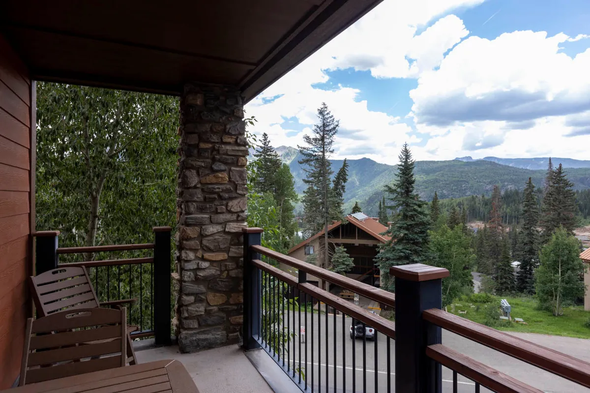 This is an exterior shot showing the condo's spacious wooden deck with sturdy stone pillars, set against a breathtaking backdrop of the San Juan Mountains. The scene evokes a tranquil atmosphere, with hints of guests relaxing on the balcony, perhaps sipping drinks or reading as the morning sun rises over the peaks.