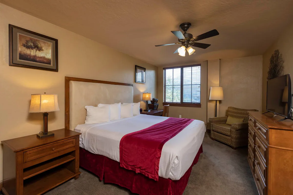 A warmly lit bedroom featuring a bed with a red accent blanket, positioned by a large window that lets in abundant natural light. The room is completed with a wooden dresser and a cozy armchair, creating a restful space ideal for winding down after a day in the mountains.
