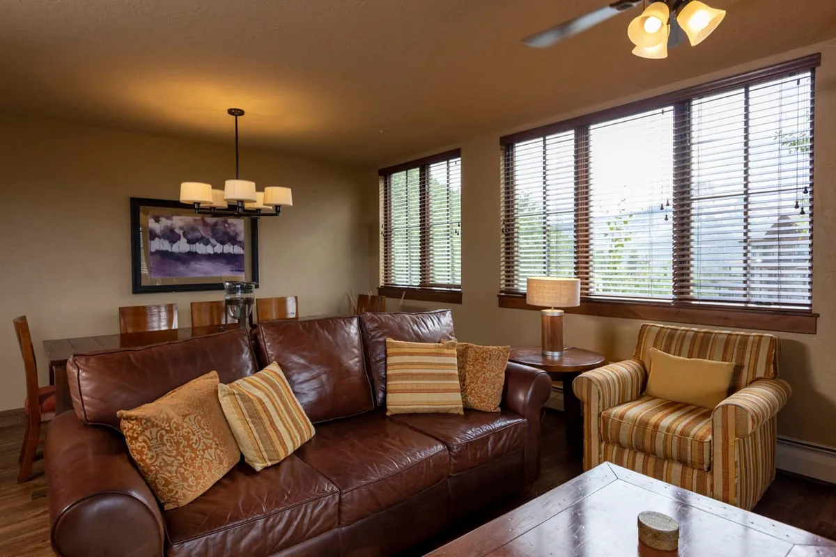 A cozy interior scene featuring a brown leather sofa and striped armchair arranged around a window with blinds, complemented by a dining table and a warm lamp. The atmosphere is inviting, suggesting relaxation after a day out and intimate evenings together.