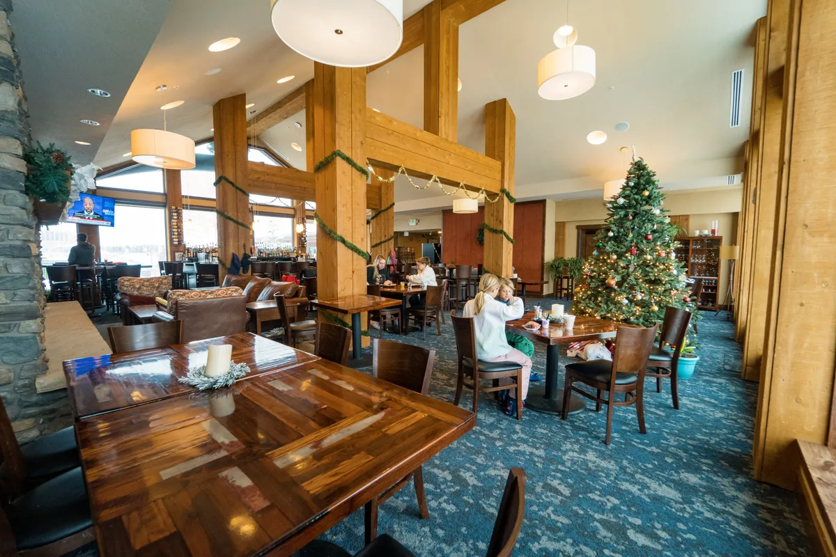 The image features a spacious, warmly lit communal area adorned for the holidays. Large wooden beams, a twinkling Christmas tree, cozy seating areas, and decorated tables invite families to gather, socialize, and enjoy celebratory meals. The atmosphere is festive and welcoming, perfect for family relaxation after outdoor adventures.