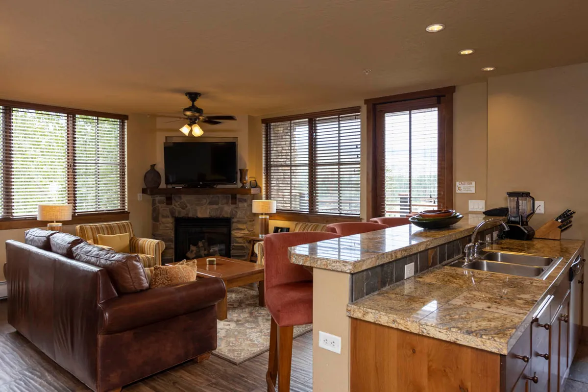 A welcoming living room featuring a prominent stone gas fireplace, plush comfortable seating, a large flat-screen TV, and expansive windows that let in natural light. The open layout leads toward a kitchen counter, reinforcing the sense of togetherness and connection in the heart of the home.