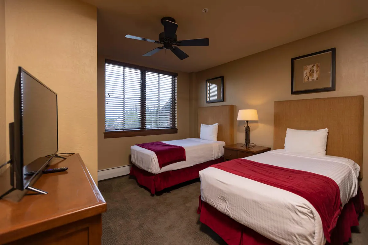 A bright bedroom featuring two twin beds with red accents, a large window with blinds, wooden furniture, and a softly lit lamp. The room is inviting, comfortable, and perfect for families or friends to unwind together.