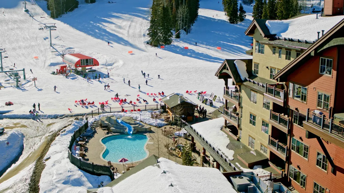 This vibrant exterior image showcases the lively ski slopes immediately adjacent to the lodge, with people skiing, snowboarding, swimming in the outdoor pool, and relaxing in the hot tub. The lodge buildings are prominent in the scene, emphasizing the ski-in/ski-out experience and resort-style amenities.