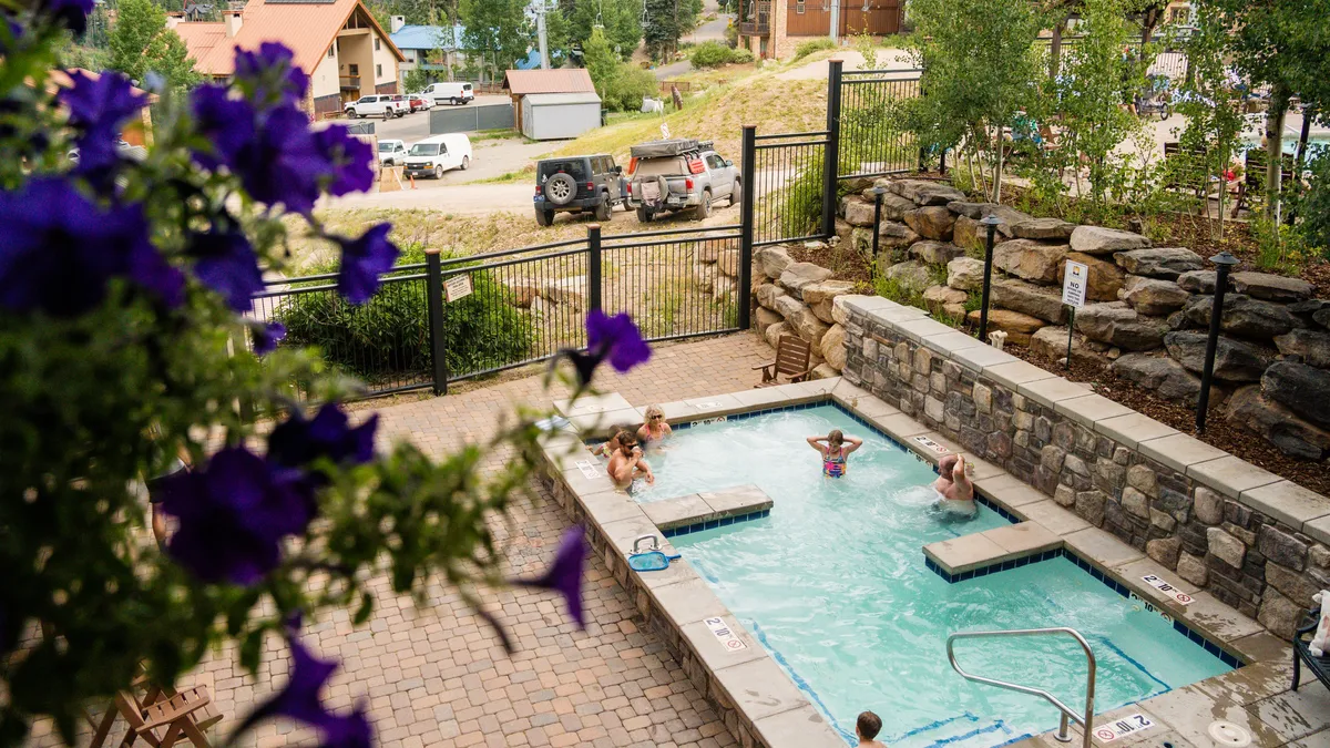A lively outdoor swimming pool scene, with people enjoying the water, surrounded by vibrant flowers and set against the backdrop of the mountains. The cheerful mood emphasizes summer relaxation and social fun.