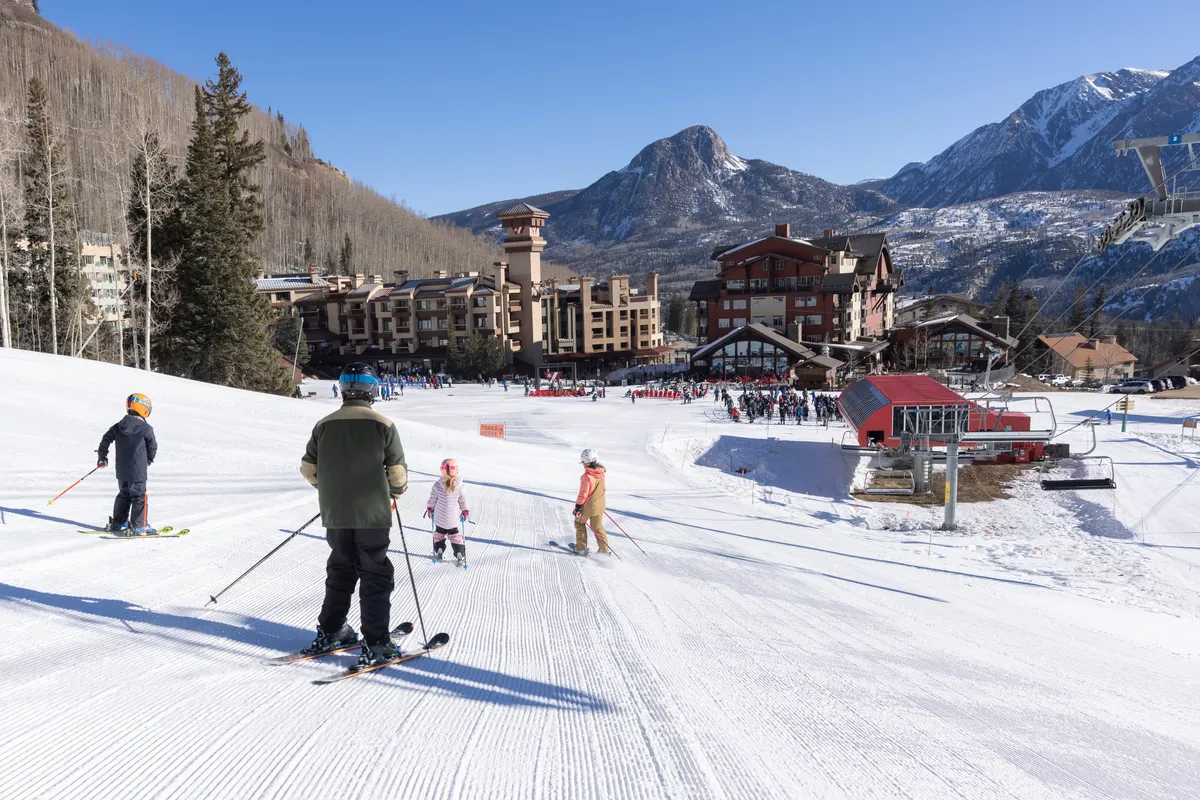 Skiers and snowboarders are captured actively enjoying the slopes in front of the resort buildings, with a ski lift and majestic mountain backdrop. The sense of motion and joy highlights the vibrancy of winter activities available at the resort.