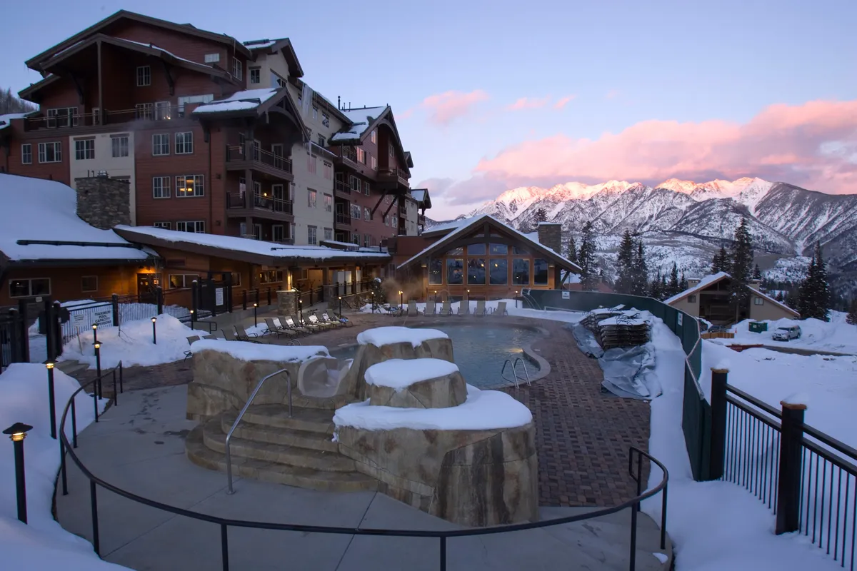 A serene view of a heated pool surrounded by snow-covered rocks, with a striking mountain backdrop and inviting architectural details of the resort. The setting is tranquil, ideal for relaxing swims or soaking in the warmth despite winter conditions.